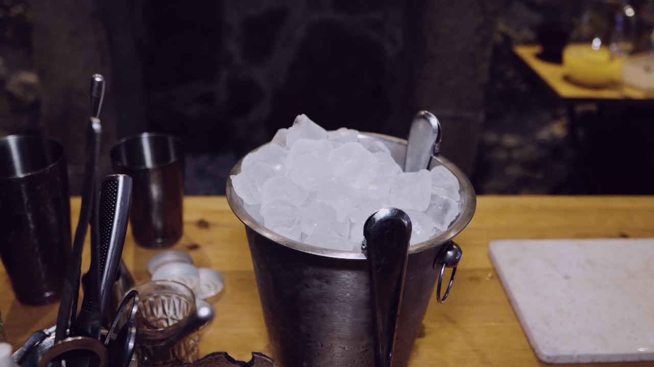 Close-up of an ice bucket with tongs on a wooden bar counter, surrounded by bar tools