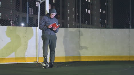 hombre con capucha y gorra apoyado contra un poste de meta metálico leyendo un libro al aire libre por la noche, una pelota de fútbol bajo los pies, una escena iluminada por las luces de la ciudad urbana