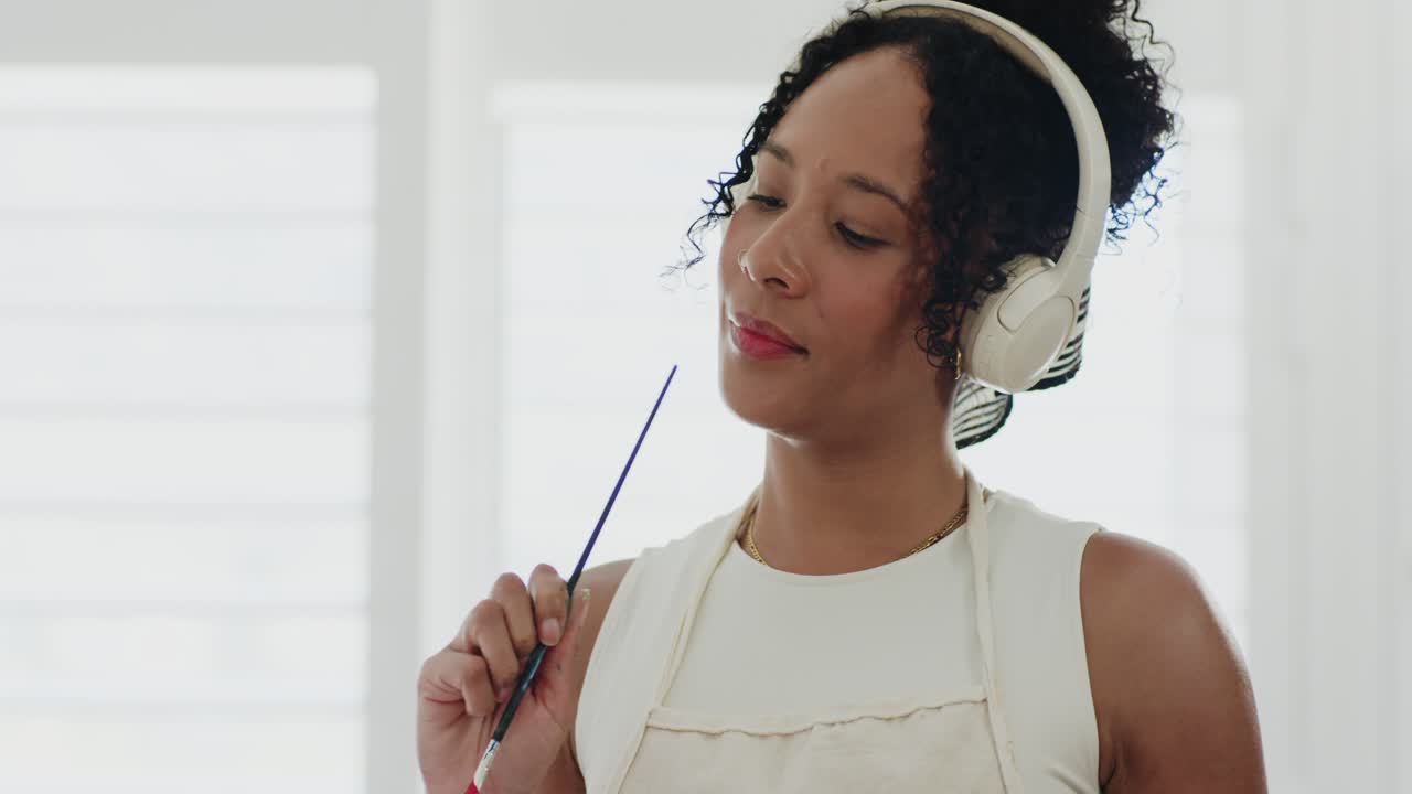 African American woman listening through headphones, tapping brush, deciding painting in studio
