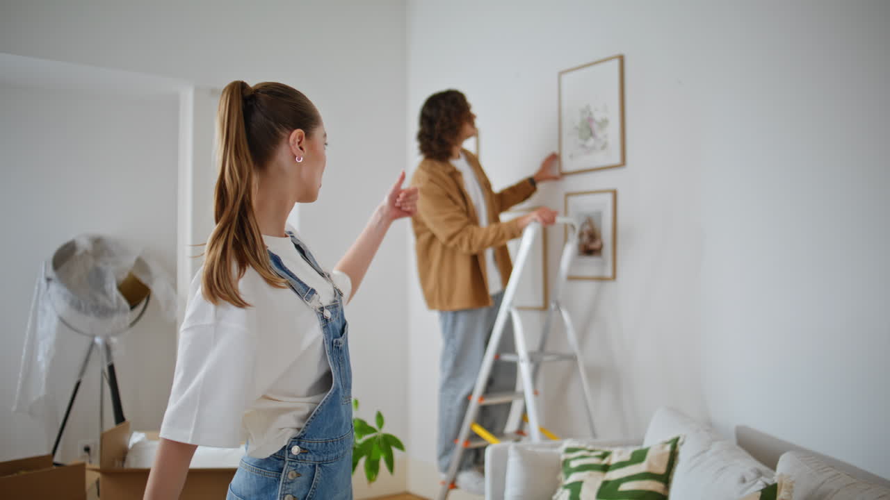 Man woman decorating wall placing artwork in living room. Husband on ladder