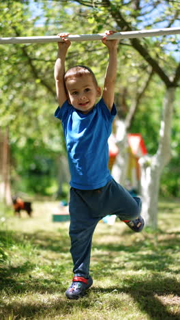 lovely kid hanging on the horizontal bar lifting his feet up. child doing sports outdoors in summer. vertical video.