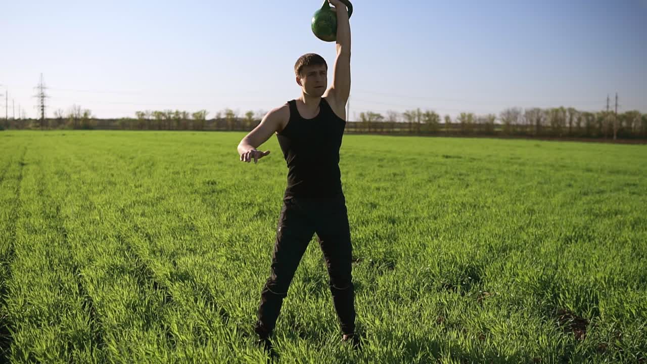 un joven con un físico deportivo levanta un peso con una mano. hermosa naturaleza, de pie en un prado verde