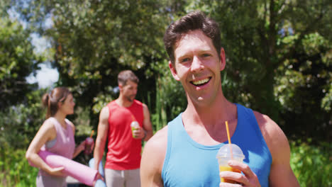 hombre caucásico sonriente bebiendo bebida para la salud, con un grupo diverso hablando después de yoga en el parque soleado