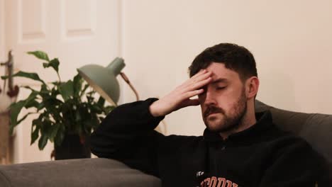 Young-man-with-beard-sat-on-couch-rubbing-head-with-a-migraine