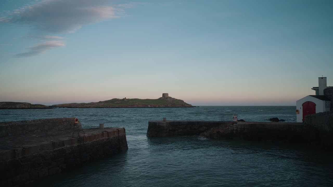 A locked-off shot of Colliemore Harbour with Dalkey Island in the background at dusk. The calm sea reflects the fading light, with the historic Martello Tower visible on the island.