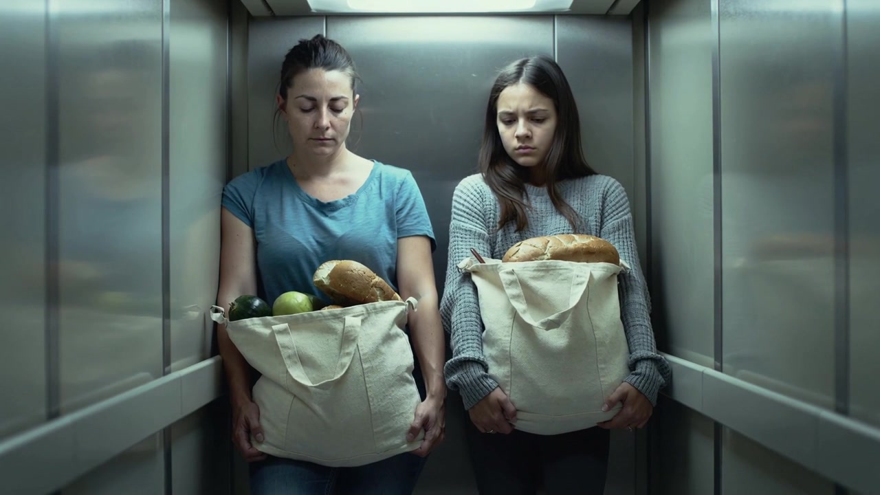 Segurando sacolas de lona com pão e frutas, mulher e adolescente em pé em elevador de metal voltando para casa