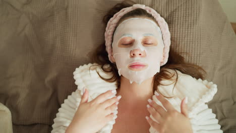 woman waves hands on face lying with on bed. brunette female in bathrobe enjoys time after spa treatments moving hands on soft bed slow motion