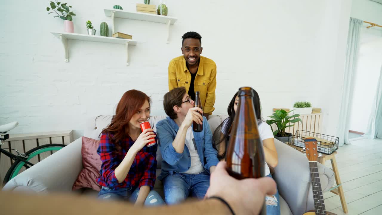 POV shot of male hand clinking beer bottle with friends while celebrating party at home indoors