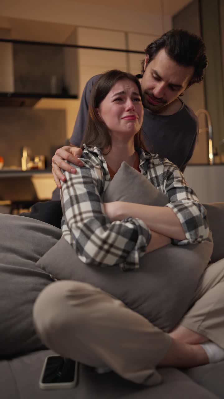 A young brunette man in a gray T-shirt calms his brunette girlfriend who is watching a very sad movie and crying while sitting on a gray sofa and hugging a pillow in a modern apartment in the evening