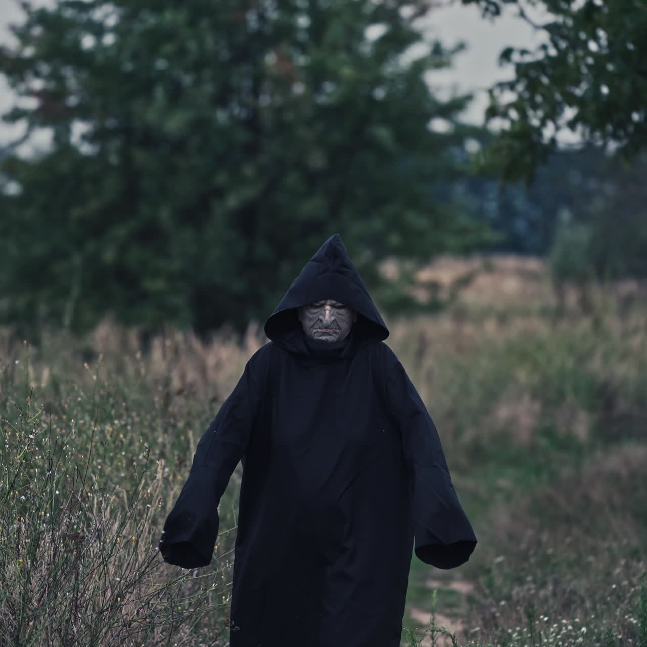 Portrait of scary death in black. Spooky figure in dark mantle with hood coming to camera on nature background. Horror concept.