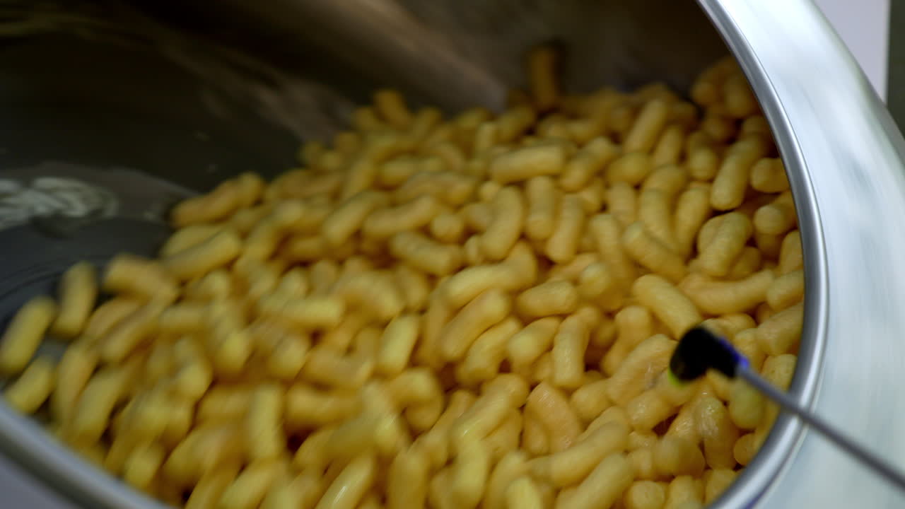 Delicious crispy snacks covered with liquid sweet syrup. Maize sticks spinning in the metal tank at food factory. Close up.