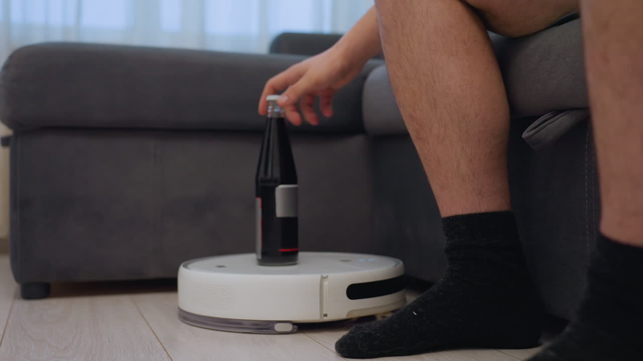 Robot vacuum transports bottle of drink across living room floor toward person sitting on sofa, creative concept of smart home automation, modern lifestyle convenience