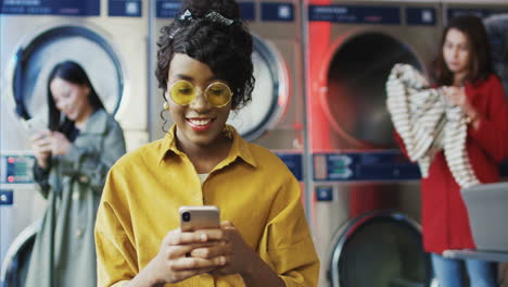 young pretty and stylish girl in yellow glasses standing in laundry service room and tapping on smartphone