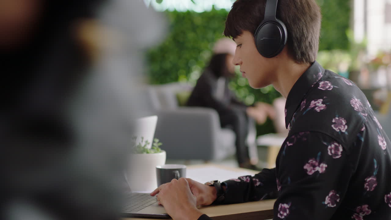 젊은 백인 남성 학생이 노트북 컴퓨터를 사용하여 창의적인 프로젝트를 수행하고 온라인 연구를 탐색하고 읽고 즐기며 음악을 듣고 트렌디한 사무실 작업 공간에서 헤드폰을 착용합니다.