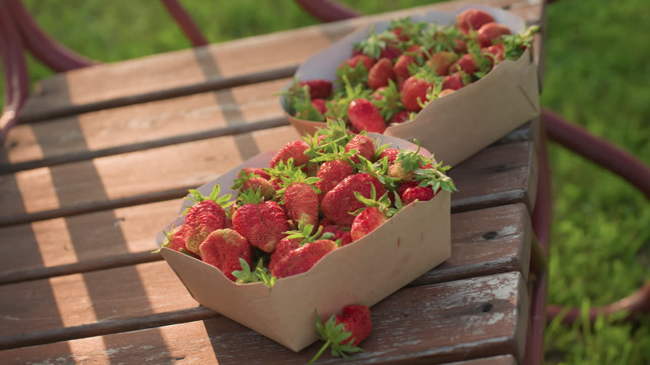close up of cardboard strawberry container on rustic swing bench gently swaying in warm golden glow someone places second container causing strawberry to tumble onto weathered wooden bench
