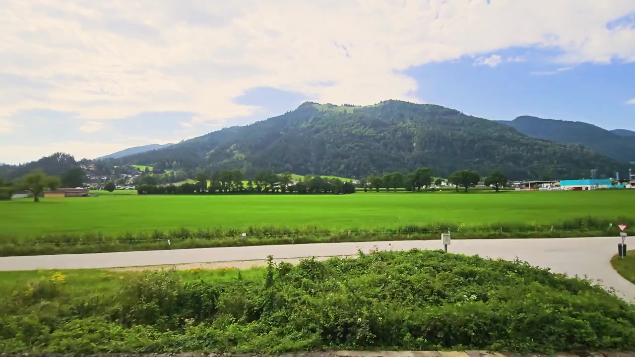 オーストリアを走行中の列車からの眺め。晴れた夏の田園風景の中、豊かな緑の牧草地、田舎の農家、アルプスの森、そして山の頂が通り過ぎる