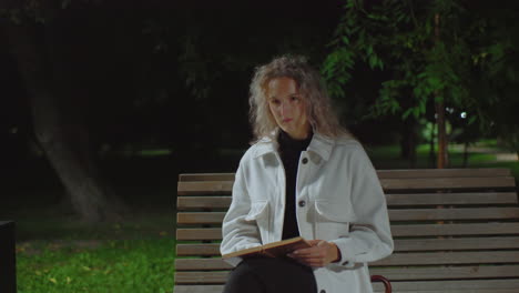 thoughtful lady seated on wooden bench at night reading book while her hair sways gently in wind, umbrella placed behind her and dim park background