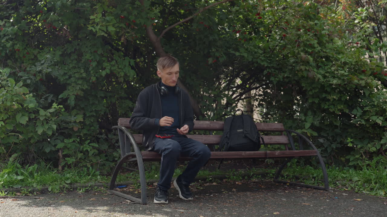 Joven con mochila sentado en un banco mirando el móvil, ajustándose los auriculares y revisando mensajes, mirando hacia un camino de grava y un seto frondoso, con luz tenue y la energía tranquila de los viajeros, sutil optimismo.