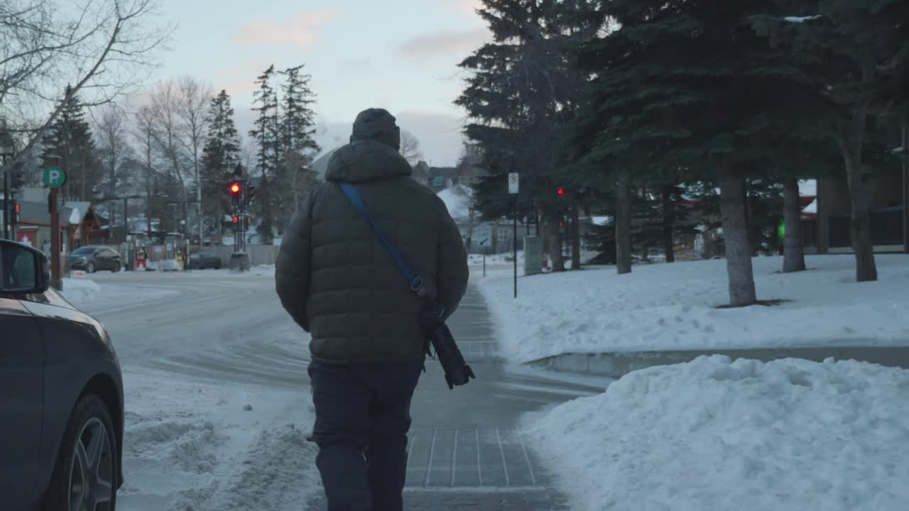 Man walking on the street during winter, with cars, trees, and buildings around.