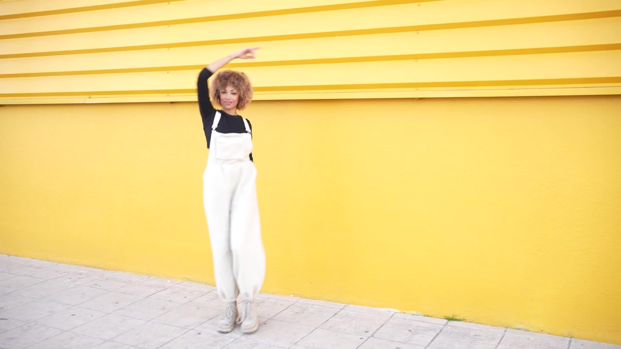 A trendy young woman enjoying dancing against a bright yellow wall while expressing joy and fun