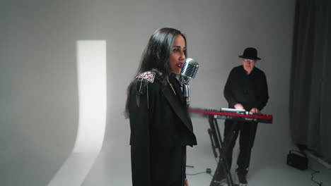 elegant vocalist in black ensemble performs at vintage microphone with keyboardist in background, white studio setting