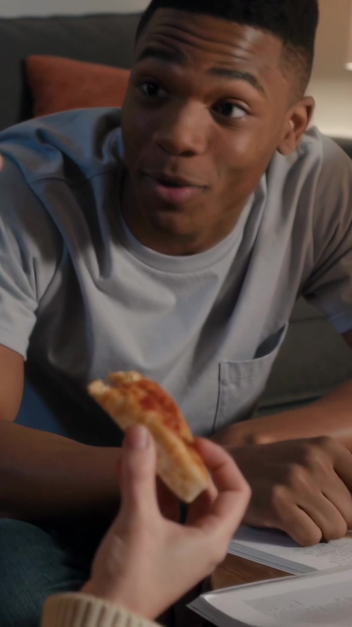 Vertical video: Taking pizza bite, woman in sweater listening while man pointing at book on sofa