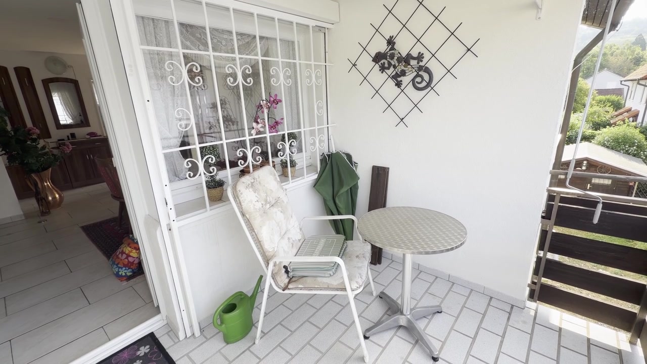 A gentle handheld shot of a bright tiled balcony featuring a round metal table, a garden chair, a folded green patio umbrella, and a window overlooking the interior of a residential home.