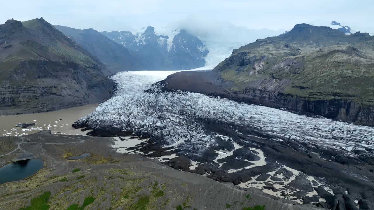 Luftdronebillede af Vatnajökull-gletsjertungen, der strækker sig gennem en vulkansk dal mod en gletsjerlagune, omgivet af forrevne islandske tinder