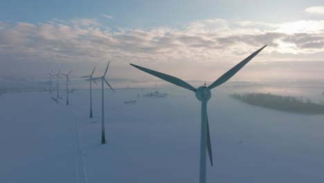 aerial establishing view of wind turbines generating renewable energy in the wind farm, snow filled countryside landscape with fog, sunny winter day, wide orbiting drone shot