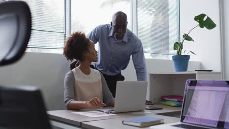 Diverse-businessman-and-businesswoman-talking,-woman-using-laptop-in-office