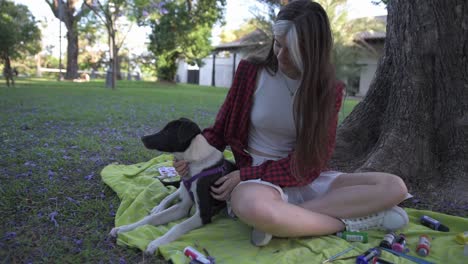 charming young woman caressing her dog sitting under tree surrounded by paintings and book at sunset