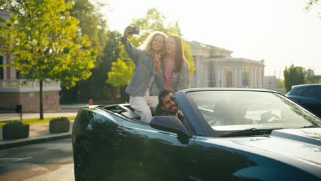 Mädchen-Sitzen-Auf-Einem-Cabrio-Und-Machen-Ein-Selfie.-Blondine-Und-Brünette-In-Jeansjacken-Machen-Ein-Selfie,-Während-Ihr-Freund-Dabei-Ist