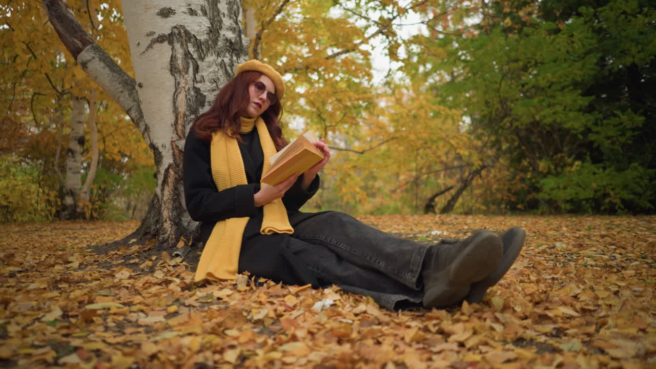 Elegante signora con basco giallo siede all'aperto, a gambe incrociate, appoggiata a un albero, leggendo un libro tra le foglie autunnali cadute, godendosi la tranquilla solitudine, circondata dalla bellezza della natura.