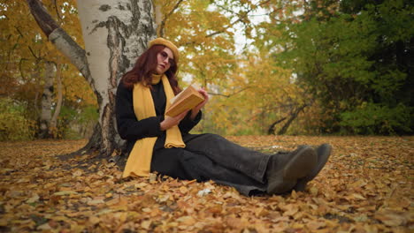 una elegante dama con boina amarilla se sienta al aire libre, con las piernas cruzadas, apoyada en un árbol, leyendo un libro en medio de las hojas de otoño caídas, disfrutando de la tranquila soledad, rodeada de la belleza de la naturaleza