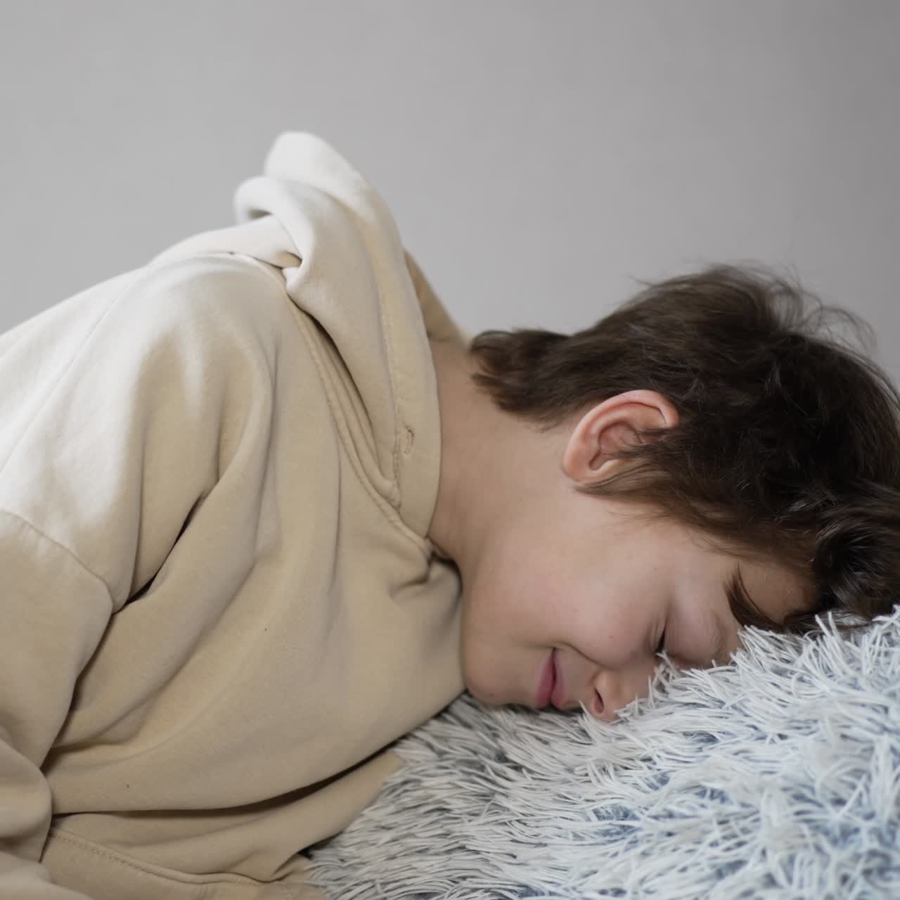 Caucasian teenager holding soft fluffy pillow in his hands. Dark-haired boy resting his face on the pillow and smiles. Grey backdrop