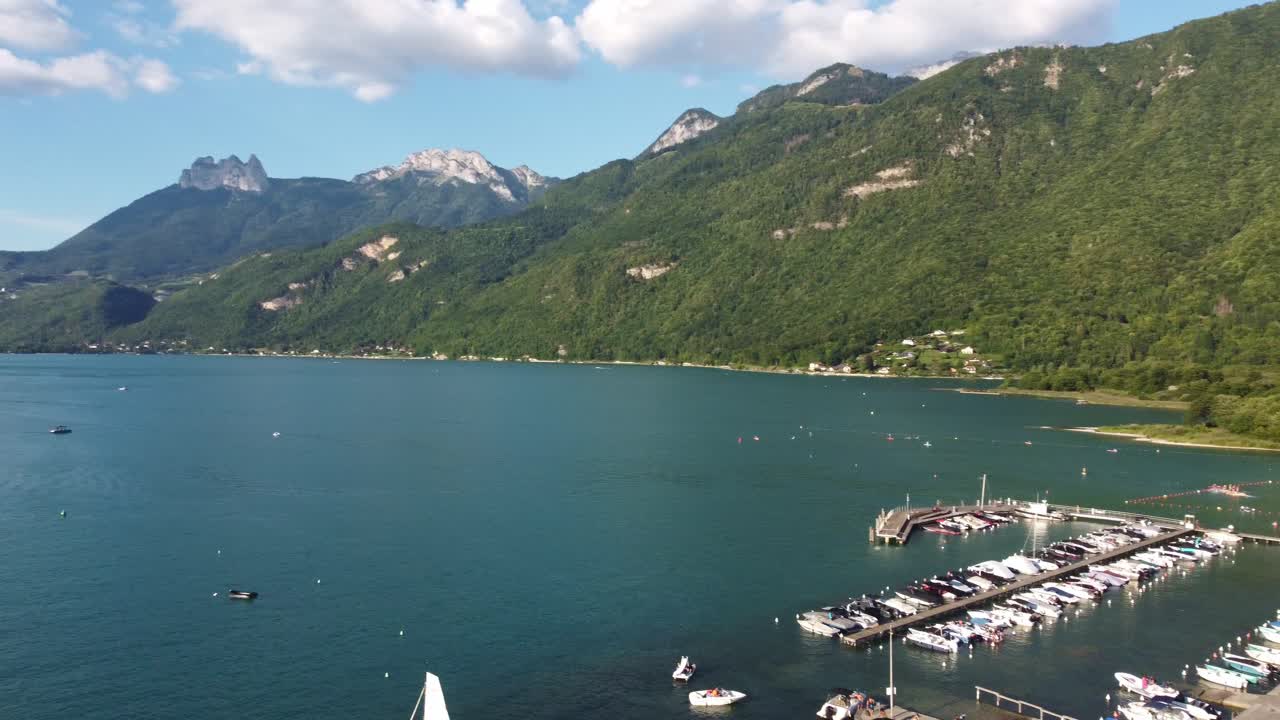 Aerial footage rises up from behind a tree to reveal a marina on Lake Annecy. Then the footage rotates left slowly to show the breath taking views of the Lake with the surrounding mountains