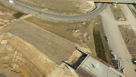 Vista-Aérea-De-La-Curvatura-De-Una-Carretera-Cuando-Se-Cruza-Con-Un-Puente,-En-Medio-De-Un-área-De-Construcción-Y-Movimientos-De-Tierras-En-Curso.