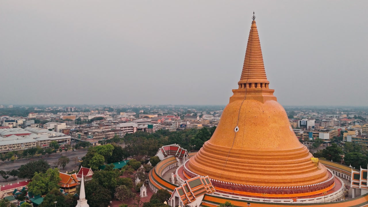 High-angle drone perspective captures giant golden stupa rising above urban center. Majestic Buddhist monument dominates city skyline in Thailand under hazy overcast sky.