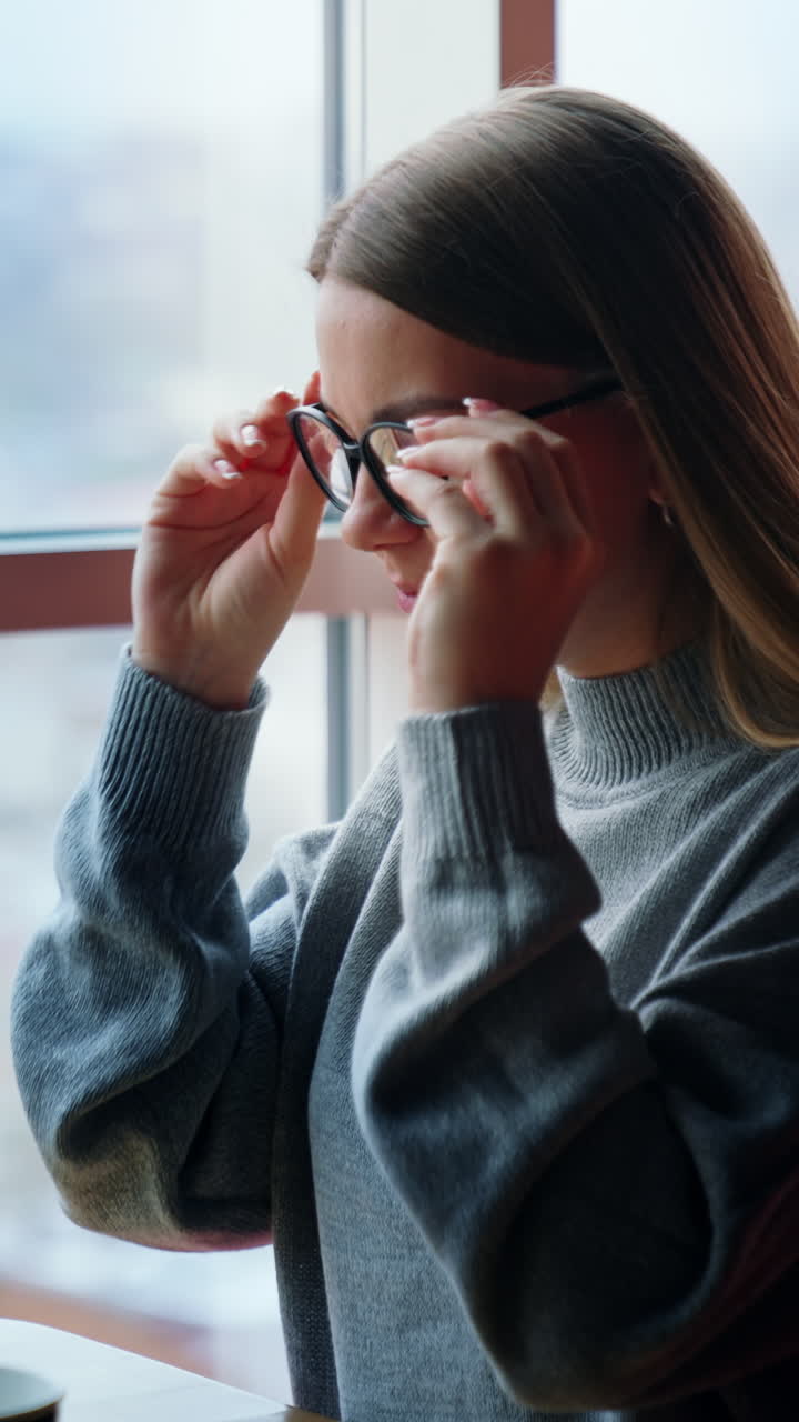 Happy positive lady puts on glasses and starts her work on laptop. Employee in a comfortable new office with panoramic window. Vertical video