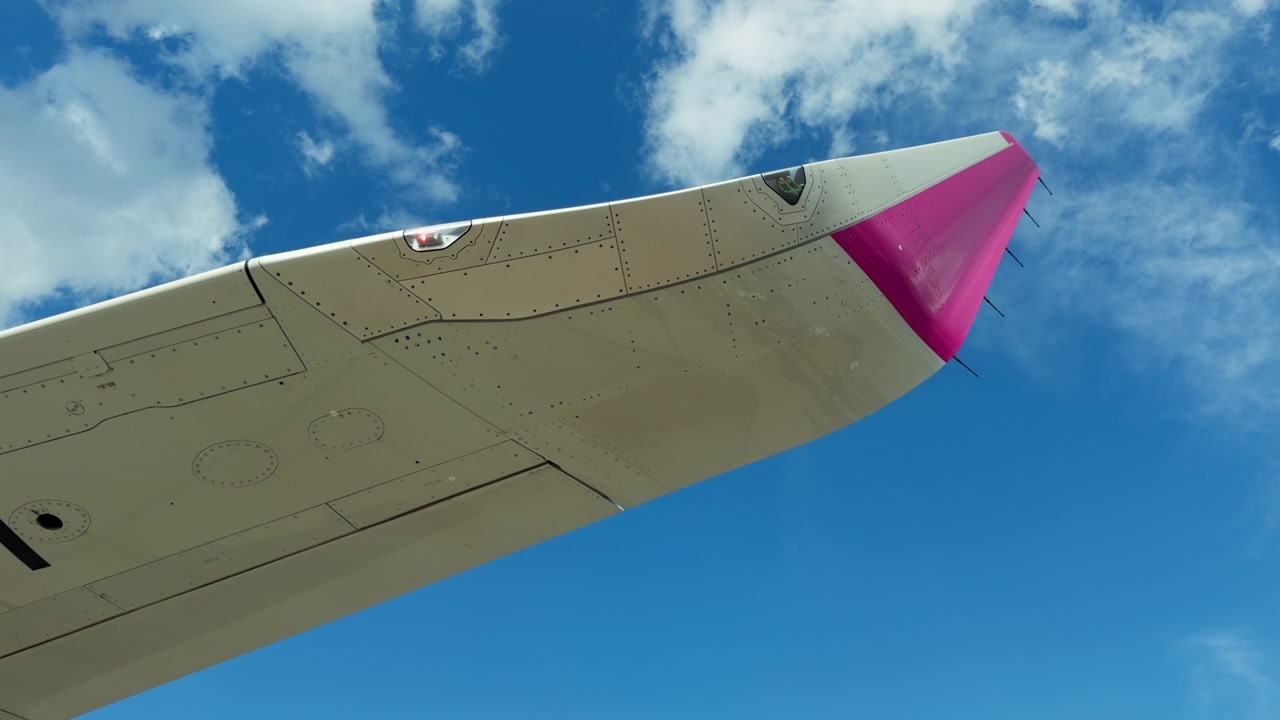 Et lavvinklet bilde viser en flyvinge med en rosa tupp. Hvite skyer krysser den blå himmelen over flyet som står parkert på bakken