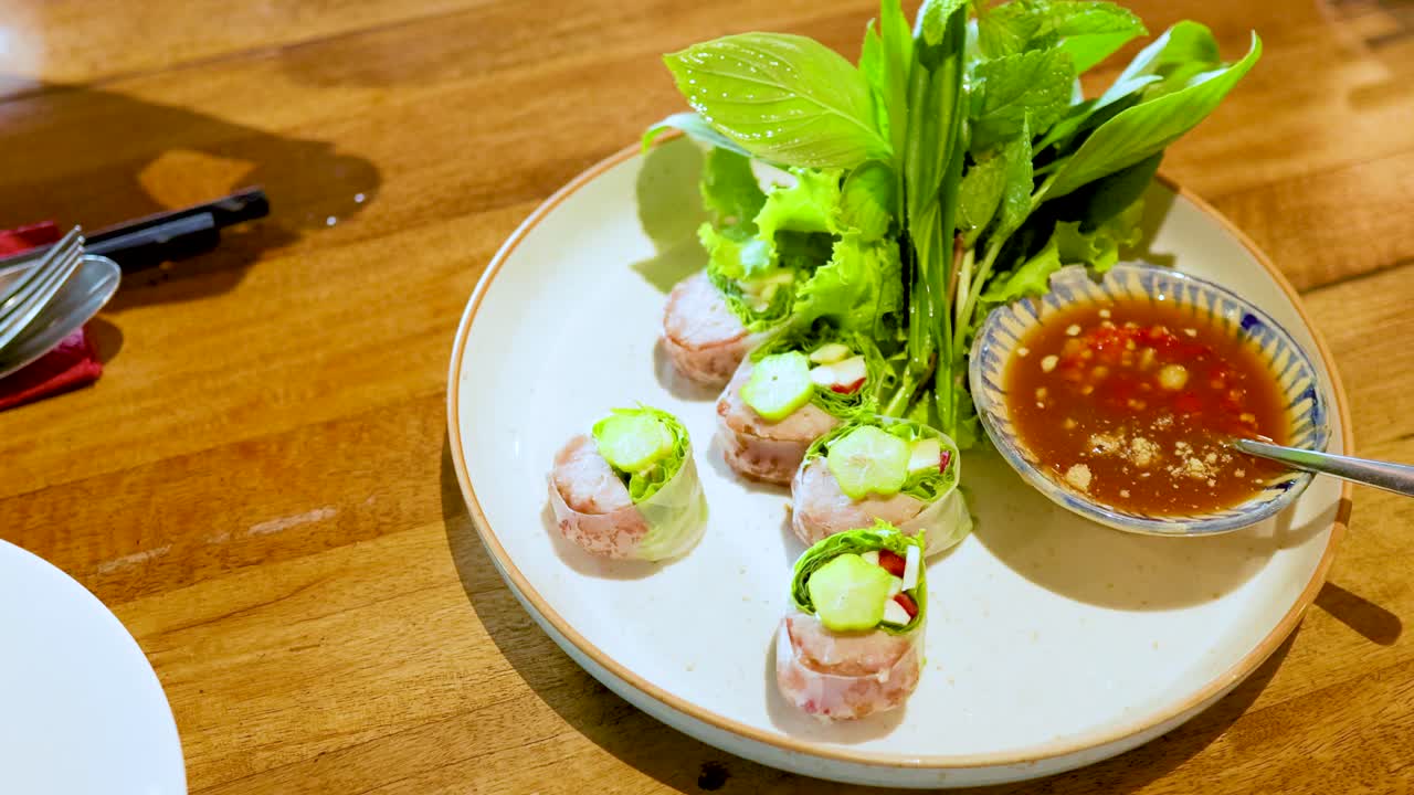Hands arrange Nem Nuong pork rolls with fresh greens and sauce on a wooden table in bright lighting