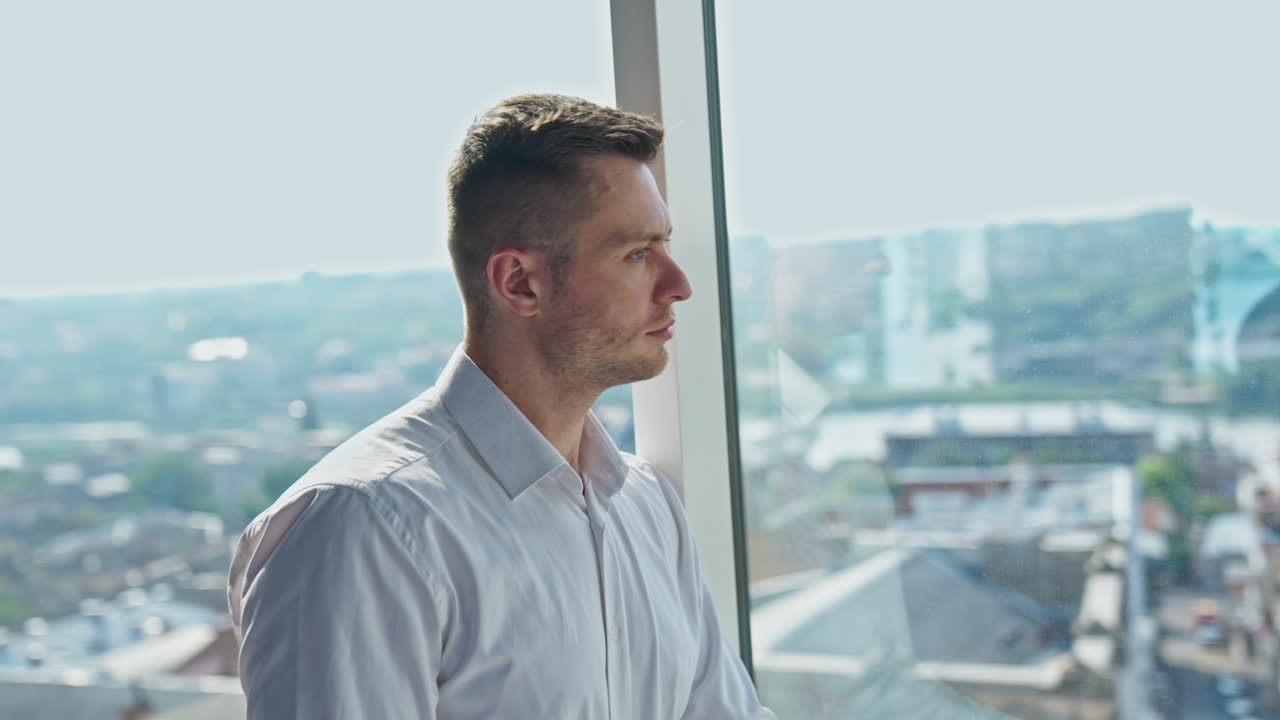 Thoughtful mid-aged man stands leaning on panoramic window. Man puts on glasses looking straight into camera smiling.