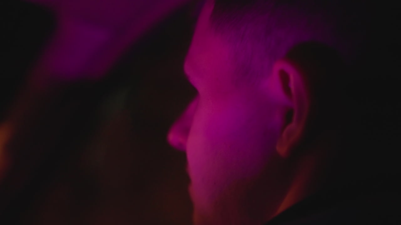 uomo bianco alla guida illuminato al neon, profilo laterale in auto con illuminazione viola magenta, finestrino riflettente ed espressione concentrata, operatore di rideshare durante il turno di notte, profondità di campo cinematografica e intima