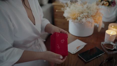 woman in white robe opening a red gift box at a beautifully decorated table with candles