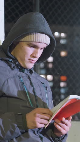 atleta con capucha y gorra apoyado contra el poste de meta metálico al aire libre por la noche, leyendo un libro pensativo bajo las luces de la ciudad urbana, un momento pacífico de reflexión, iluminado por las luces callejeras