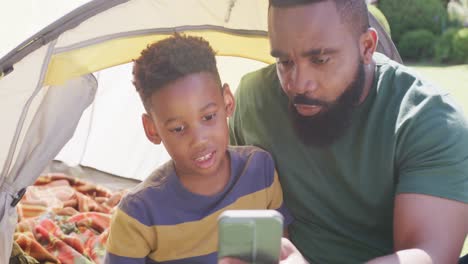 happy african american father and son sitting in tent and using smartphone, in slow motion