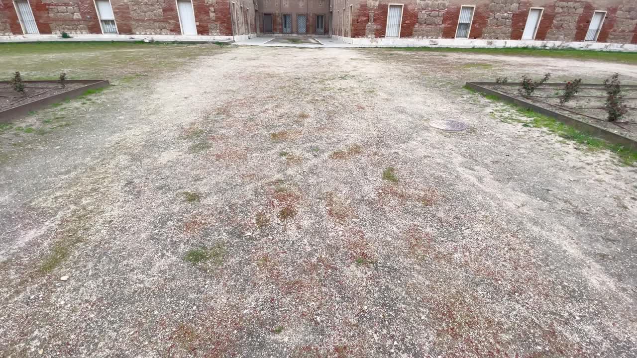We see a white dirt road and with a camera elevation we discover one of the internal patios of the House of Trades in Aranjuez with its stone and old brick decoration in the Herreriano style.