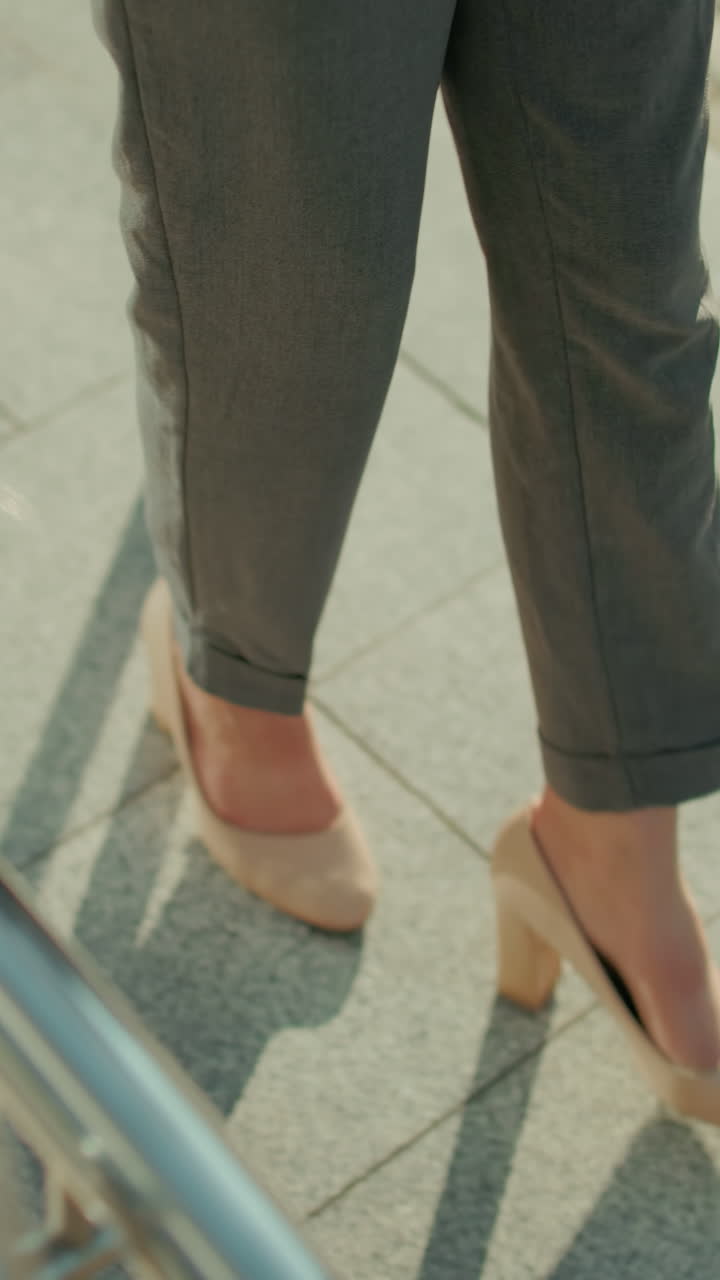 Top down view of lady in heels and blazer walking along tiled pavement bordered with iron railing in bright daylight, showcasing confident stride and urban style in professional outdoor environment