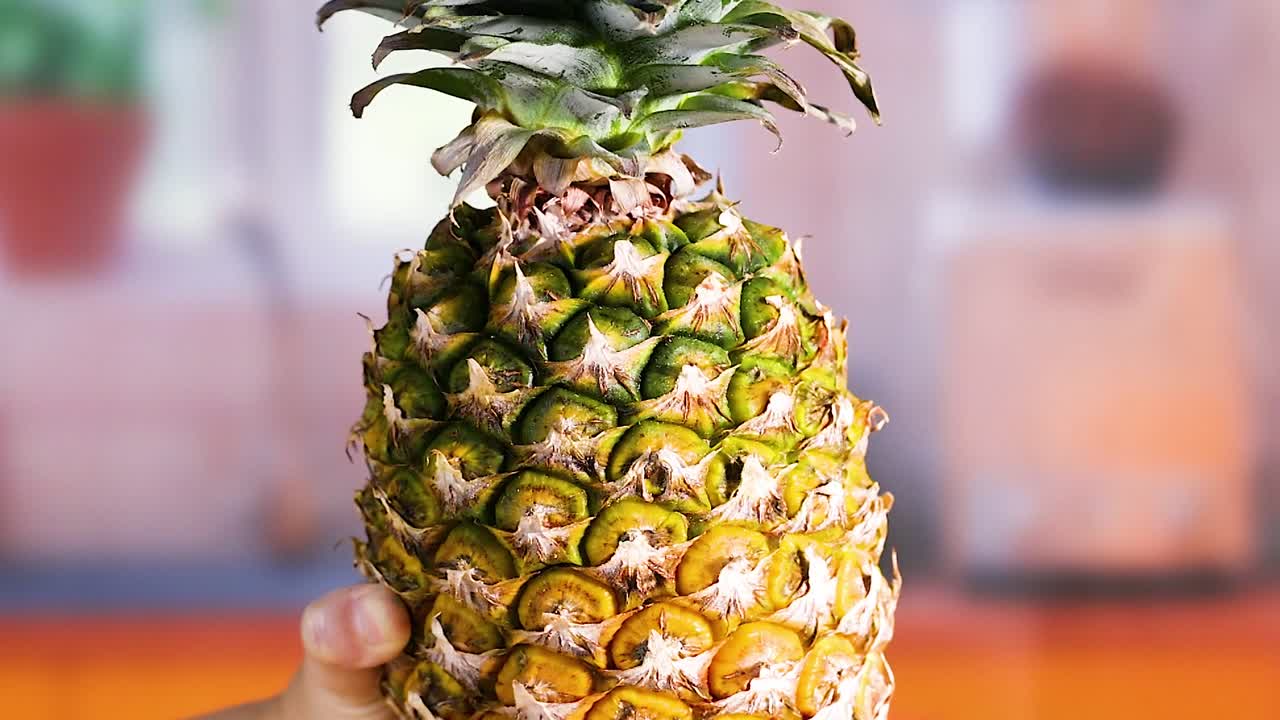 A person's hands hold and rotate a whole ripe pineapple against a blurred kitchen background