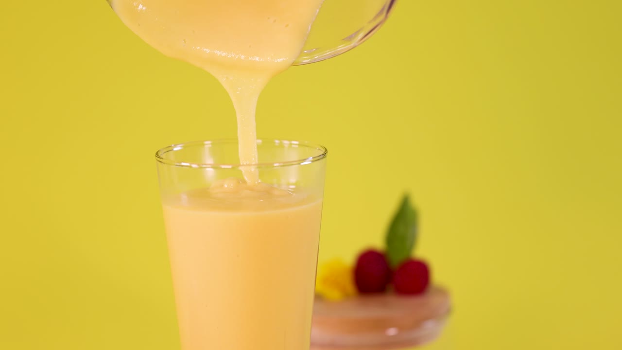 Creamy mango smoothie poured into glass, vibrant yellow background, bright lighting, minimal camera movement
